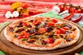 Italian Pizza with hunting sausages, tomatoes, chili peppers, cheese and pickles on a round wooden board