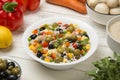 Italian rice salad in a white porcelain salad bowl on a white wooden table with ingredients.