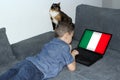 Italy flag on a laptop display, a little schoolboy in jeans lies on a sofa and scrolls, a cat sits nearby, a concept of studying