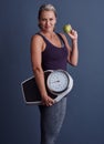 Its a lifestyle. Studio portrait of an attractive mature woman holding an apple and a weightscale against a blue Royalty Free Stock Photo