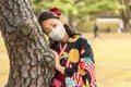 Woman in kimono wearing a mask leaned against the trunk of a pine tree.