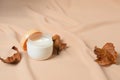 A jar of cream for dry skin care with dried autumn leaves on a wavy beige background made of chiffon