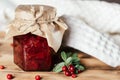 Jar of homemade lingonberry and pear jam with craft paper on lid on wooden table next to fresh lingonberries and towel