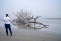 Woman photographs a washed up driftwood tree on Driftwood Beach during a very foggy,