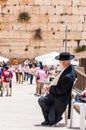 Jewish orthodox in black suit and hat sitting near entrance alley to the Western Wall. People, locals and tourists walking around
