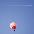 Jet airplane with long white trail flies over a hot air balloon in a beautiful blue sky Royalty Free Stock Photo