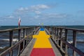 A jetty with a colourful carpet in blue, yellow and red. A blue ocean and sky in the background