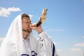 Jewish man in tallit blowing shofar outdoors. Rosh Hashanah celebration