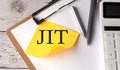 JIT word on a yellow sticky with calculator, pen and clipboard