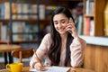 Job hunting. Happy arab woman talking by smartphone with hr representetive and taking notes, sitting at cafe interior Royalty Free Stock Photo