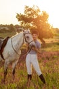 Jockey young girl petting and hugging white horse in evening sunset. Sun flare