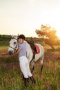 Jockey young girl petting and hugging white horse in evening sunset. Sun flare