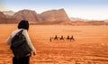 Jordan, Wadi Rum. Caravan of camels with drovers in the desert, rock mountains at sunset. Tourist from behind looks at the