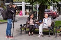 Journalist interviews an elderly man sitting on a bench in a city park, cameraman filming it on sunny summer day.