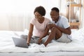 Joyful african couple sitting on bed, watching movie on laptop