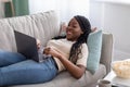 Joyful african lady laying on couch at home, using laptop