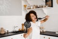 Joyful African young woman having fun with kitchen utensils. African american housewife in apron singing with spatulas Royalty Free Stock Photo