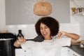 Joyful African young woman having fun with kitchen utensils. African american housewife in apron singing with spatulas Royalty Free Stock Photo