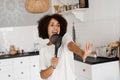 Joyful African young woman having fun with kitchen utensils. African american housewife in apron singing with spatulas Royalty Free Stock Photo