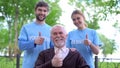 Joyful aged pensioner and two volunteers showing thumbs up, social support fund
