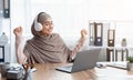 Joyful arabic woman in hijab listening music in headphones at workplace