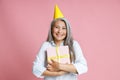 Joyful Asian lady with hoary hair and bright party hat holds gift box in studio