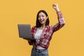 Joyful asian lady holding laptop and gesturing yes, shaking clenched fist, celebrating success and great news