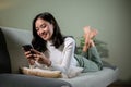 A joyful Asian woman is enjoying chatting with her friends on her phone while laying on a couch