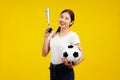 Joyful Asian woman holding a soccer ball and cheer horn isolated on yellow background Royalty Free Stock Photo