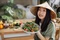 Joyful asian woman in traditional conical hat holding healthy salad from organic vegetables