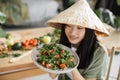 Joyful asian woman in traditional conical hat holding healthy salad from organic vegetables
