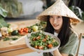Joyful asian woman in traditional conical hat holding healthy salad from organic vegetables
