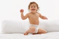 Joyful baby, little girl with curly hair smiling and sitting on white bed with pillow behind against white studio Royalty Free Stock Photo