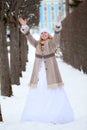 Joyful beautiful bride in coat and wedding dress stands in winter park and rising hands up Royalty Free Stock Photo