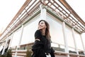 Joyful beautiful young woman with a cute smile in a vintage coat in a trendy black t-shirt in stylish white jeans posing in a city Royalty Free Stock Photo