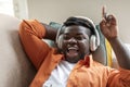 Joyful black guy reclining on sofa, singing songs, using headphones Royalty Free Stock Photo