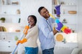 Joyful black man and his girlfriend singing during housecleaning, using mop and duster as microphones