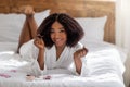 Joyful black woman lying on bed in robe, applying nail polish, making manicure, smiling at camera Royalty Free Stock Photo