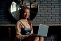 A joyful black woman working on a laptop at the coffee-shop with a black brick wall. Remote work, remote office concept.