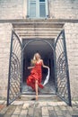 Joyful blonde jumping in red sultry dress, feeling happy. Old building, European city. Summer holiday, fashion, lifestyle concept