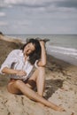 Joyful boho girl in white shirt sitting on sunny beach. Carefree stylish woman smiling and relaxing on seashore. Summer vacation.