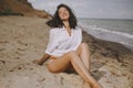 Joyful boho girl in white shirt sitting on sunny beach. Carefree stylish woman smiling and relaxing on seashore. Summer vacation.