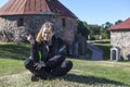 Joyful Caucasian woman in motorcycle jacket, knee guards sitting on green meadow in some landmark, copyspace