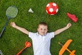 A joyful child lies on the grass with a scooter, a racket, a shuttlecock, a ball, a sneaker, and an airplane lying around him. The