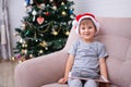 Joyful child wears santa hatlooking at the camera at christmas while sitting in a chair in the living room at home, holding a tabl