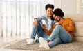 A joyful couple sitting on the floor against a couch, sharing a light-hearted moment with a smartphone