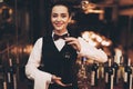 Joyful elegant waitress holding bottle of red wine, standing near bar.
