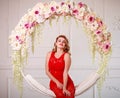 Joyful elegant woman in red dress on swing in flowers