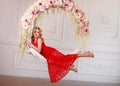 Joyful elegant woman in red dress on swing in flowers
