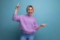 joyful energetic blond young woman in a purple hoodie shows her hands up and to the side on a blue background with copy